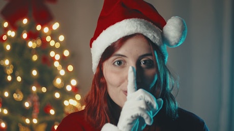 Girl makes quiet sign with hand next to Christmas tree.