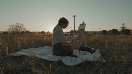 Girl paints a picture of the countryside on a canvas at sunset.