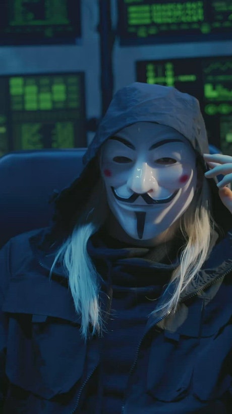 Hacker in a scary mask sitting in front of computer screens.