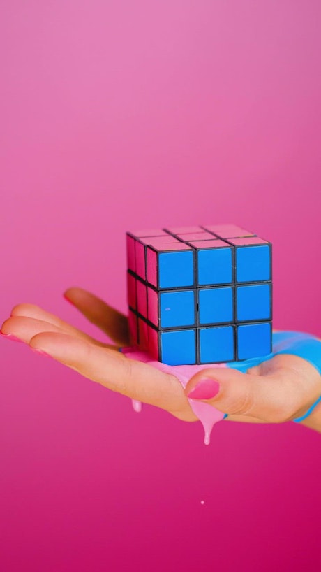 Hand holding a rubik cube that seems to melt on a pink background.