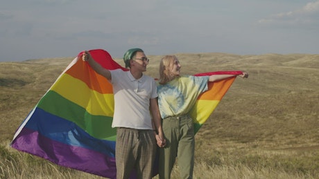 Happy couple holds a Pride flag on a meadow in the sun.