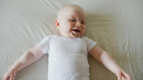 Laughing baby lying on bed in white onesie.