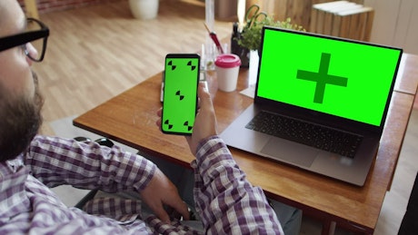 Man holding a phone with a green screen in front of a laptop.