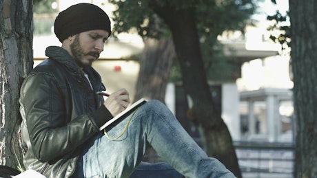 Man in a beanie writing in a book in public.