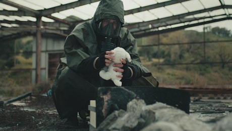 Man in gas mask finds a teddy bear in a destroyed warehouse.