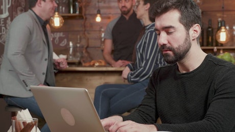 Man pauses to think while typing on laptop in cafe.