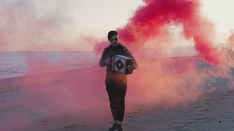 Man plays music with accordion on the beach against red smoke.