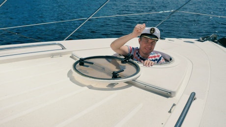 Man wearing a captains hat emerges from the porthole of a yacht.