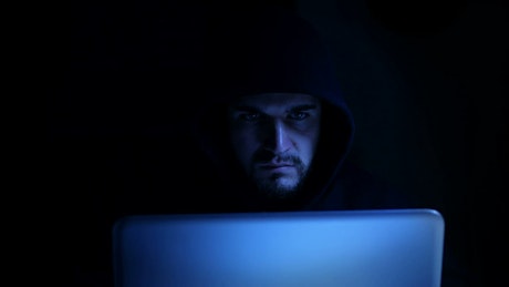 Man wearing a hoodie in a dark room typing aggressively on a laptop.