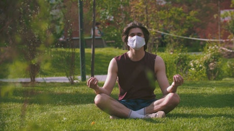 Man with a face mask meditating in a sunny park.