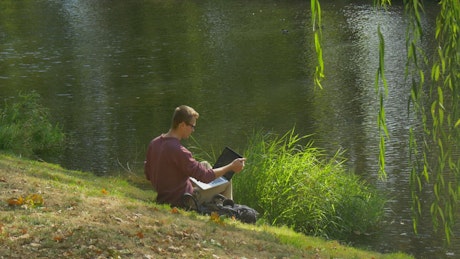 Man with a laptop on the bank of a river.