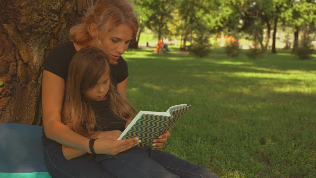 Mother and daughter read a book together in a park.