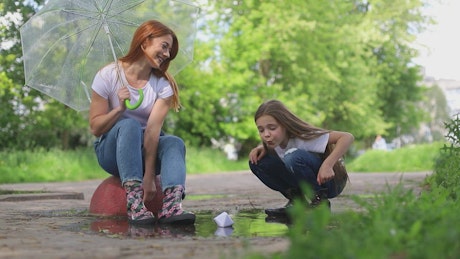 Mother and teenager play with paper boat in a puddle of water.