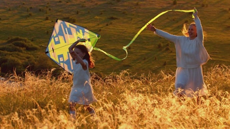 Mother with her daughter running on the hill with a kite at sunset.