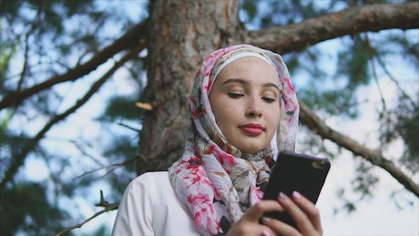 Muslim girl wearing a head scarf checking her cell phone at the park.