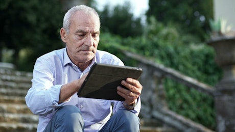Old man sitting on steps using a digital tablet.