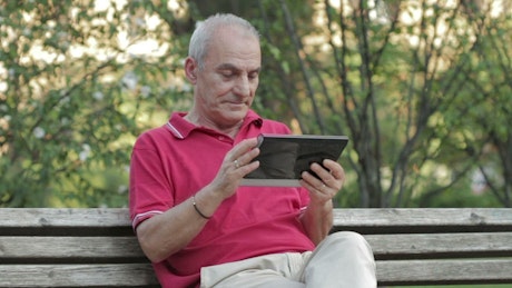 Older man sitting on a bench in the park using a digital tablet.