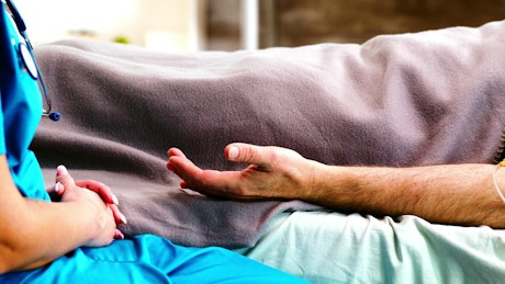 Sick man on bed comforted by nurse holding hands.