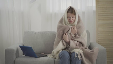 Sick woman sneezes on sofa while wrapped in blanket