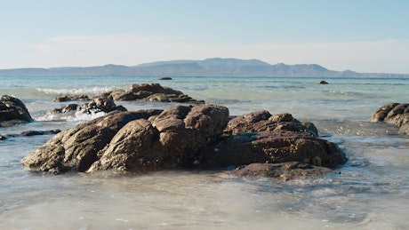 Small waves crashing on wet rocks near the shore with majestic mountains in the background.