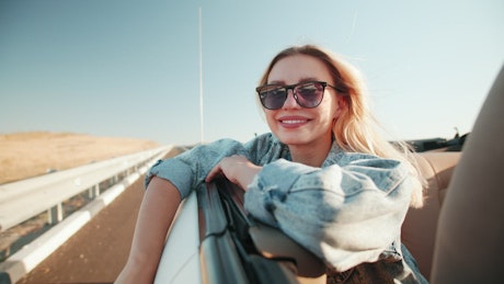 Smiling blonde woman enjoys the wind from a convertible car.
