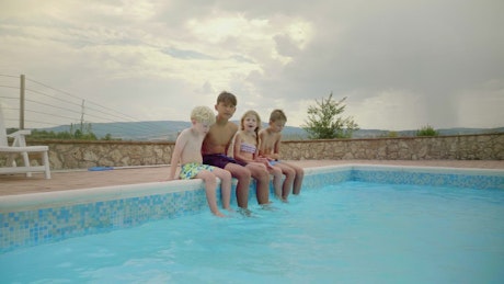 Smiling kids sit on poolside with feet in water