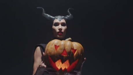Spook horned witch holds up carved pumpkin.
