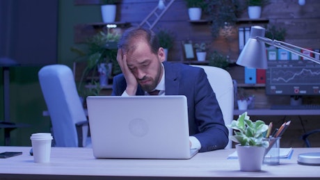 Stressed businessman rubs eyes while working on laptop.