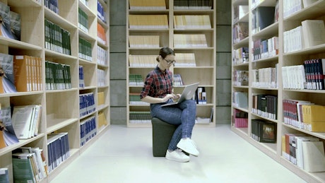Student in a library on a laptop between shelves.