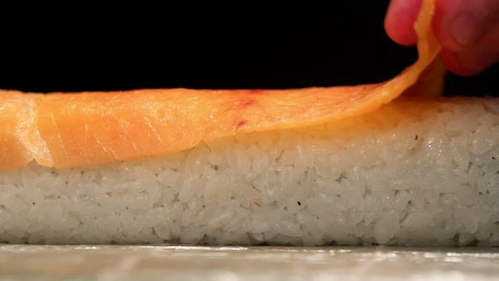 Thing piece of salmon being laid atop a sushi roll.