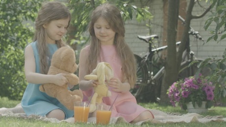 Twin girls play with teddy bear on blanket outside.