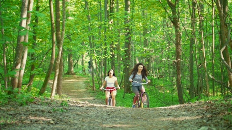 Two girls happily cycling through a forest.