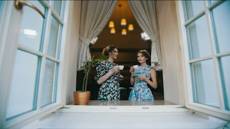 Two women drinking tea and talking at the window.
