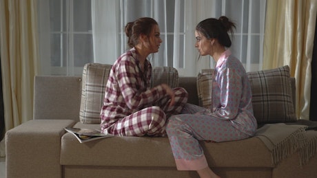 Two young women gossip in pajamas on sofa.