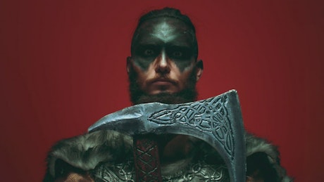 Viking in war paint holds battle axe in front of red background.