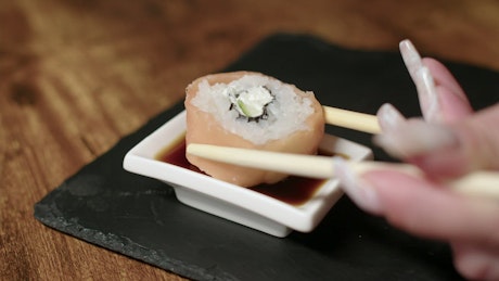 Woman dipping a piece of sushi into soy sauce with chopsticks.
