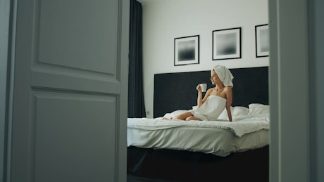 Woman enjoys coffee on bed after shower.