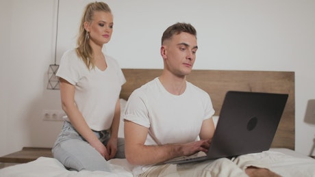 Woman hugs man working on laptop in bed