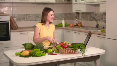 Woman prepares dinner while video chatting on laptop.