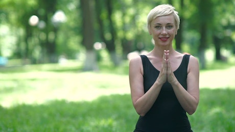 Woman smiles in namaste yoga pose in park.