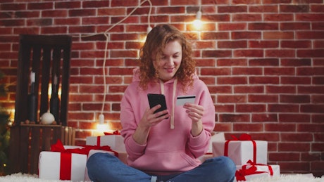 Woman surrounded by Christmas presents making a payment on her phone.