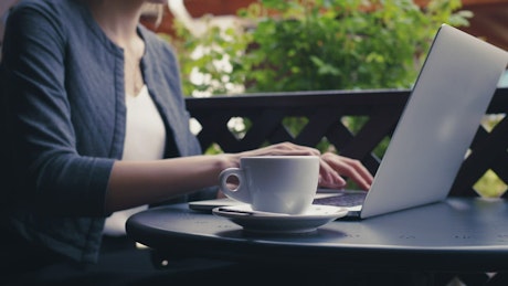 Woman typing on a laptop as she sips from a coffee cup.