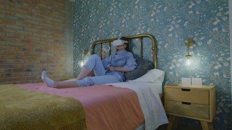 Woman using a virtual reality headset sitting on her bed.