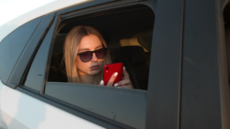 Woman wearing sunglasses scrolls on phone in the back of a taxi.