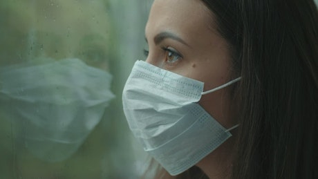 Woman with mask looking at rain through window.