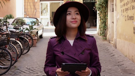 Young Asian woman walks along street using a tablet.
