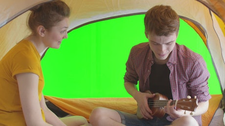Young couple sitting in a tent while camping playing the ukulele.