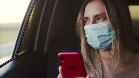 Young woman wearing a face mask checks phone int he back of a taxi.