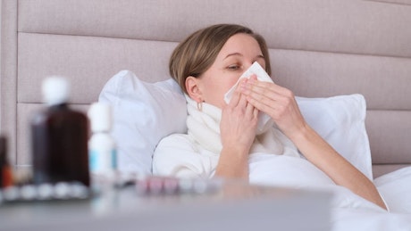 Young woman with flu blows her nose while lying in bed.