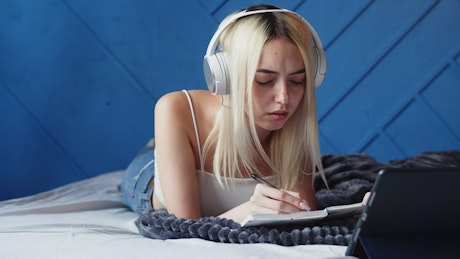 Young woman with headphones writing in her diary in bed.